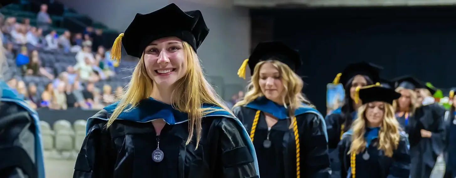 Group of graduates in caps and gowns walking down aisle at commencement ceremony.